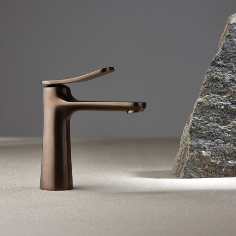 Eine schlichte, moderne Bronze-Armatur steht auf einer glatten, beigen Oberfläche neben einem strukturierten Stein. Ein neutraler grauer Hintergrund und eine sanfte Beleuchtung unterstreichen das elegante Design.